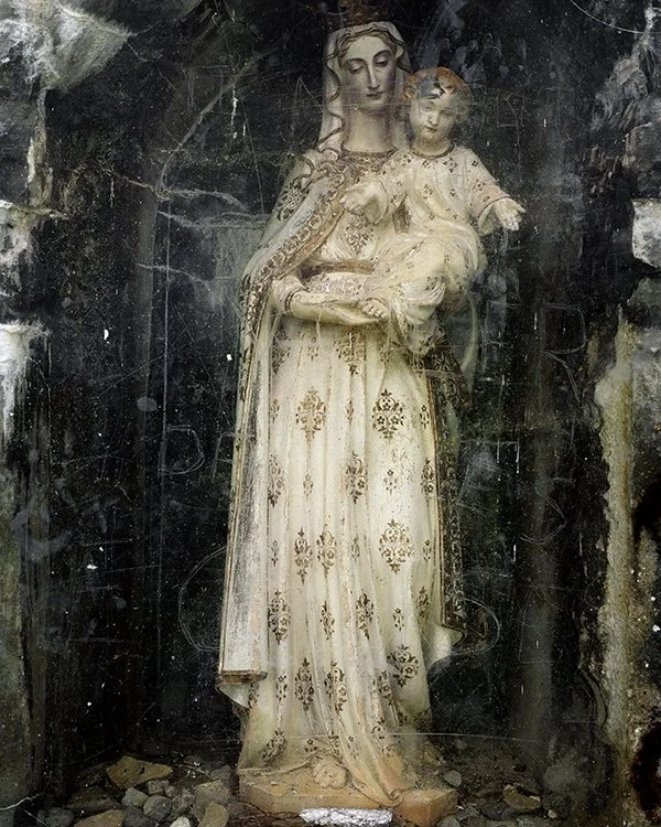 Statue de la Vierge Marie tenant l'enfant Jésus, en bois peint, dans un environnement sombre et abîmé.