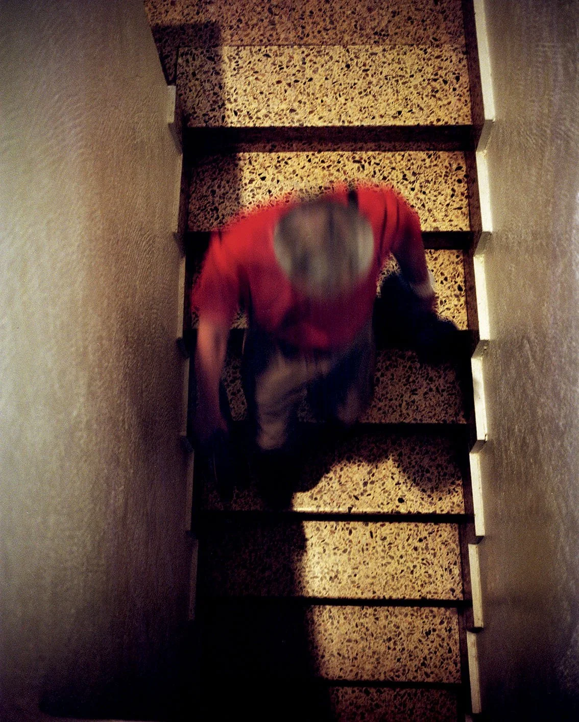A man wearing a grey hat, red shirt and beige pants descends the stairs into a narrow alley, seen from above.