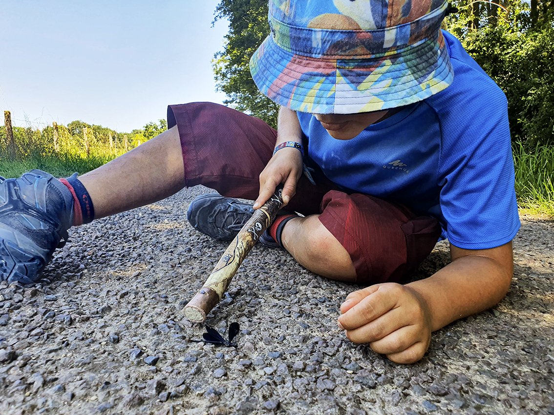 A child lying on a gravel path, wearing a colorful hat, blue T-shirt, burgundy shorts and holding a walking stick, in a natural outdoor setting.