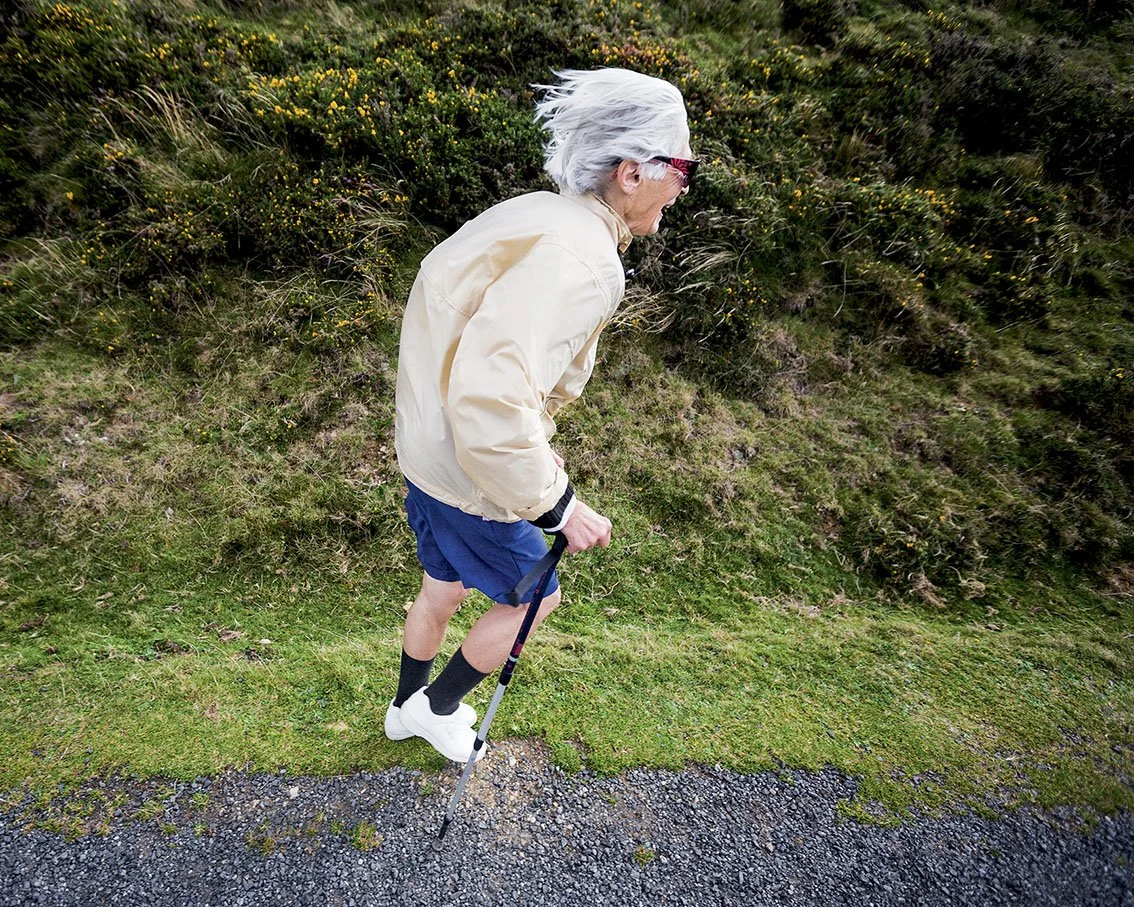 An old man Nordic walking on a country path, wearing a beige jacket, blue shorts, white shoes, black socks and using a cane.