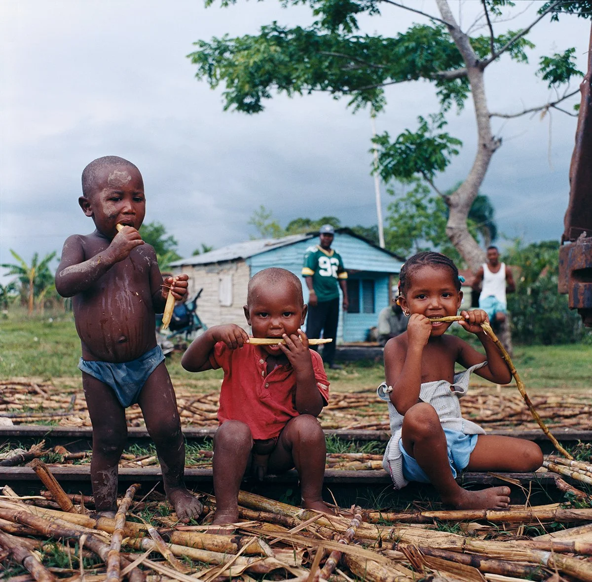 Three native children play with sticks in a rural area with a wooden house in the background and a cloudy sky.