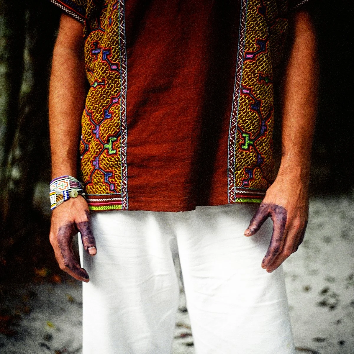 A person wearing a colorful garment with a traditional motif, a red shirt and white pants, in the middle of nature.