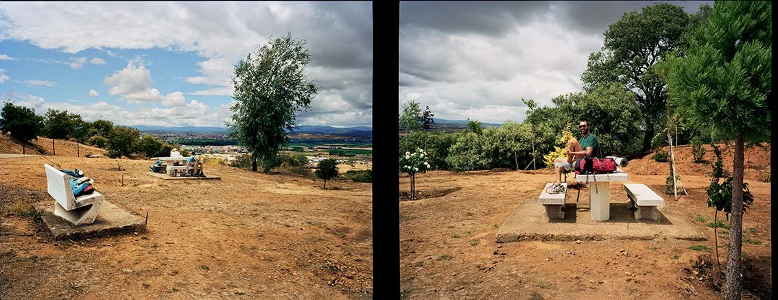 Two stone benches with backpacks and clothes on them, one person sitting opposite, in an open-air setting with trees and a cloudy sky.