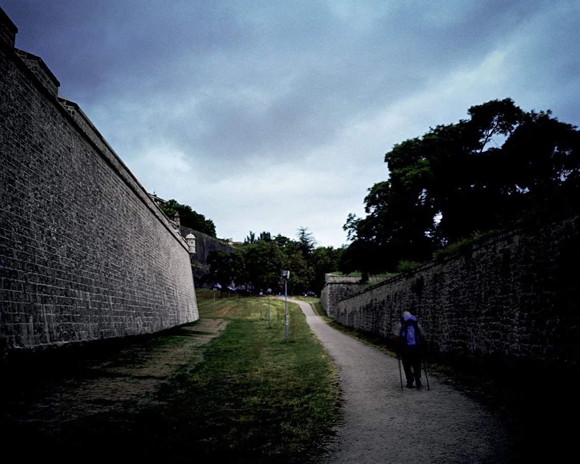 Path lined with high stone walls with an elderly person carrying a rucksack and a stick, under a cloudy sky.
