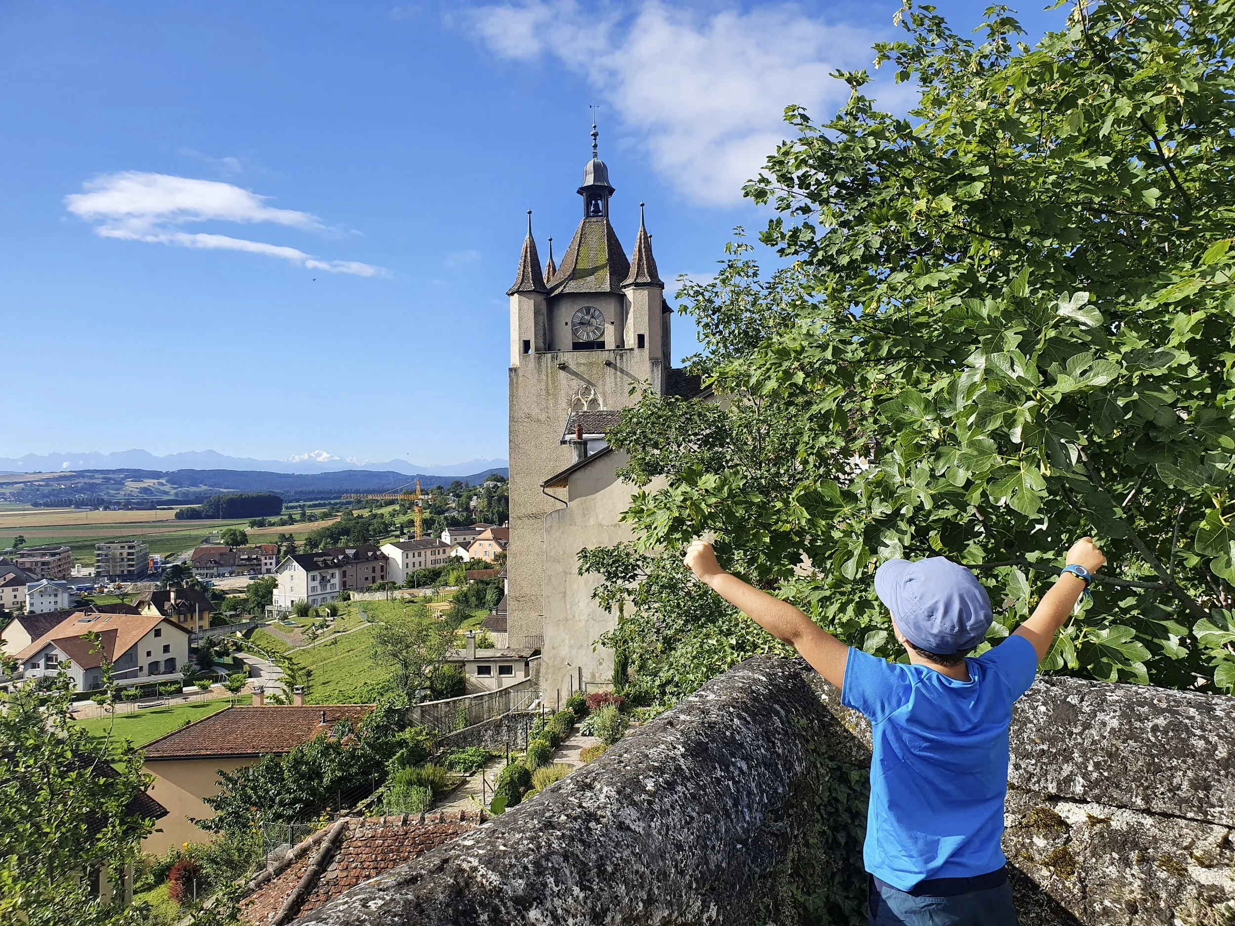 A child in a blue T-shirt and blue hat, with arms raised, looks up at an old church tower in a rural landscape with houses, fields and distant mountains on a sunny day.