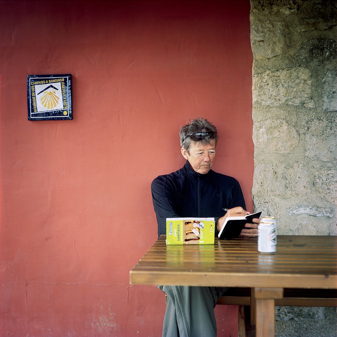 Woman seated at a wooden table, using a tablet, with a canned drink in front. Red wall background with a stone corner and a poster of the pilgrimage route to Santiago de Compostela.