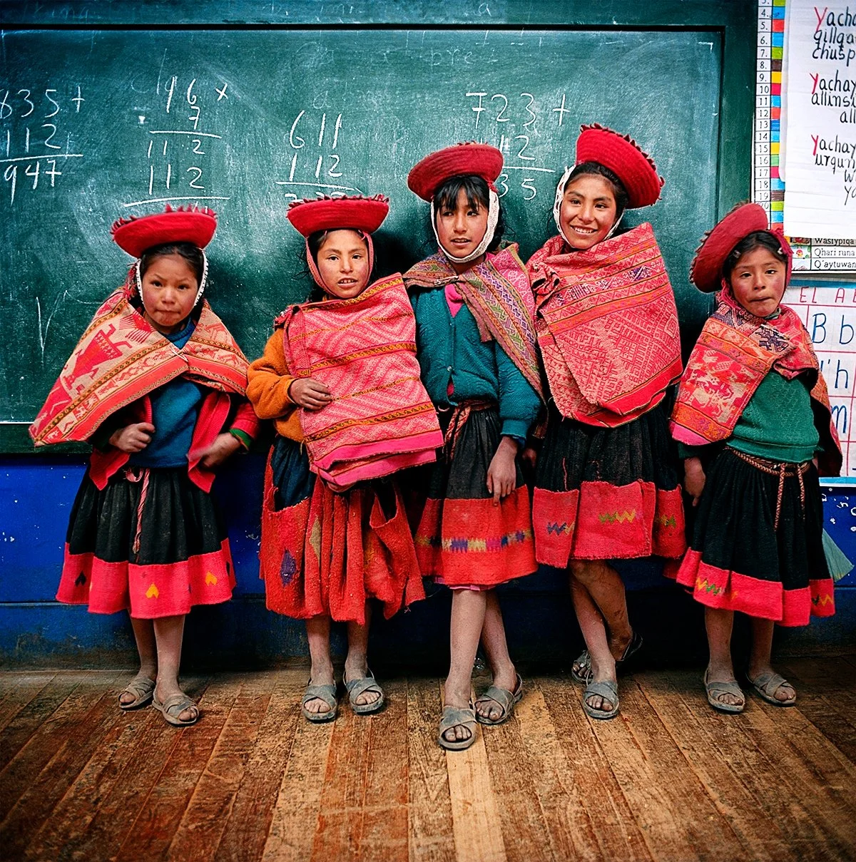 Five Andean children dressed in colorful traditional costumes, wearing red hats, in a classroom in front of a blackboard with mathematical equations written on it.