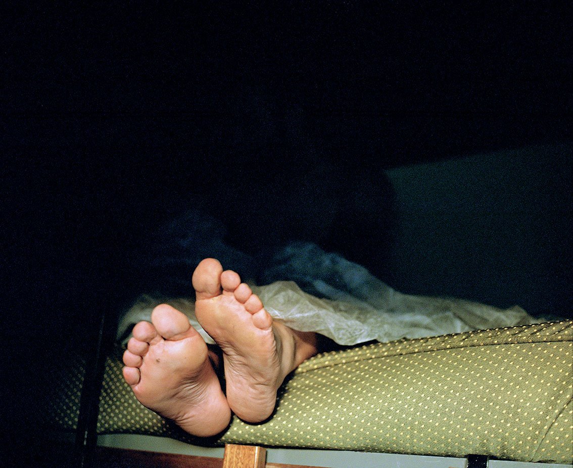 Bare feet of a person lying under a sheet, seen from the heels and soles, on a surface with a green cushion with white dots.