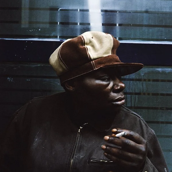 Homme noir portant un chapeau beige et marron, une veste en cuir, tenant une cigarette, assis près d'une vitre ou d'une surface en verre.