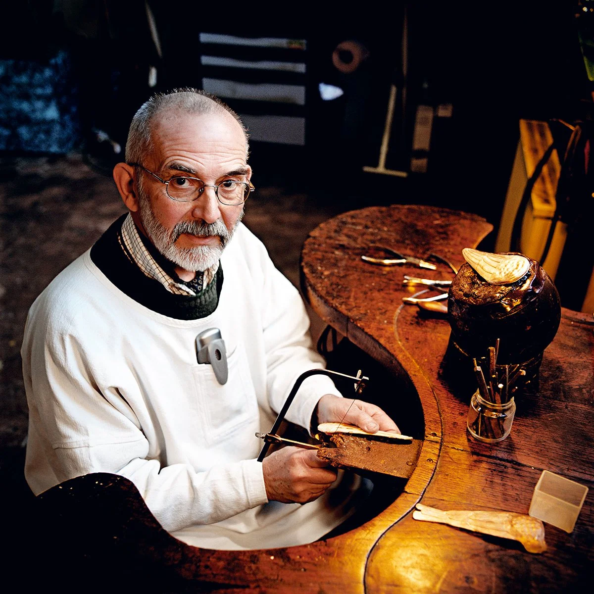 An elderly man with glasses, a beard and a serious expression, working on an artistic achievement with a hacksaw on a wooden countertop. The environment is dark, with tools and a shell-shaped work of art vi