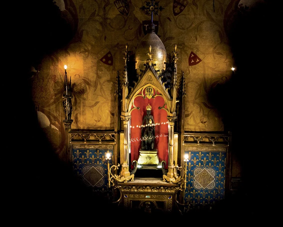 A religious sculpture of a saint or sacred figure in a chapel, framed by an ornate altarpiece, illuminated by small lights, in a wall decoration with Gothic motifs.