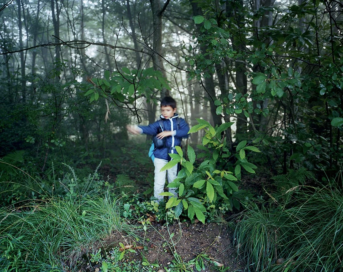 A boy in a blue jacket and backpack explores a dense green forest.