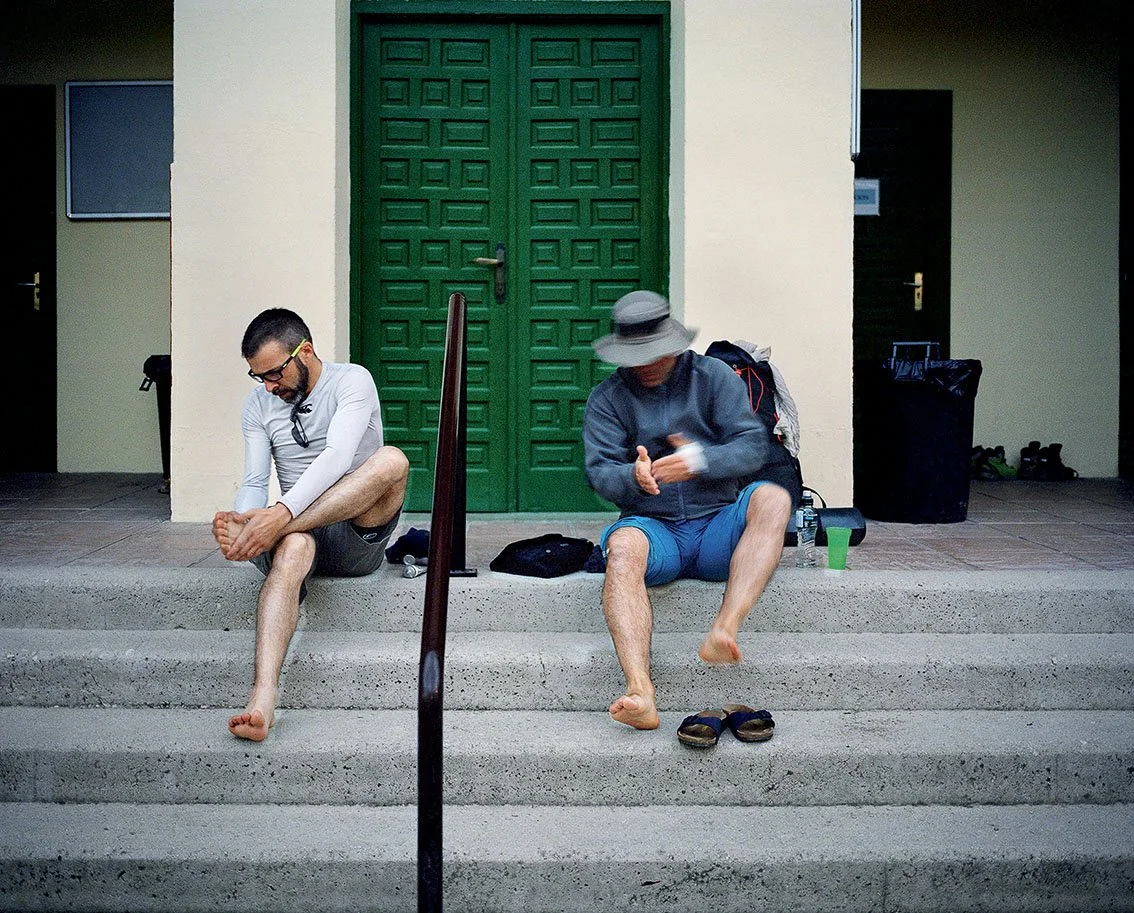 Two men sitting on steps in front of a green door, one adjusting his ankle and the other looking at his phone, with bags and a bottle of water beside them.