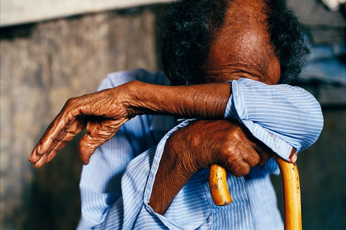 An elderly woman rests with a cane, her head buried in her crossed arm.