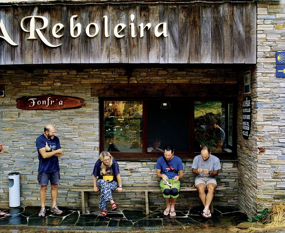 Group of people sitting and standing in front of a stone building with a sign saying "A Reboleira" in a relaxed atmosphere.