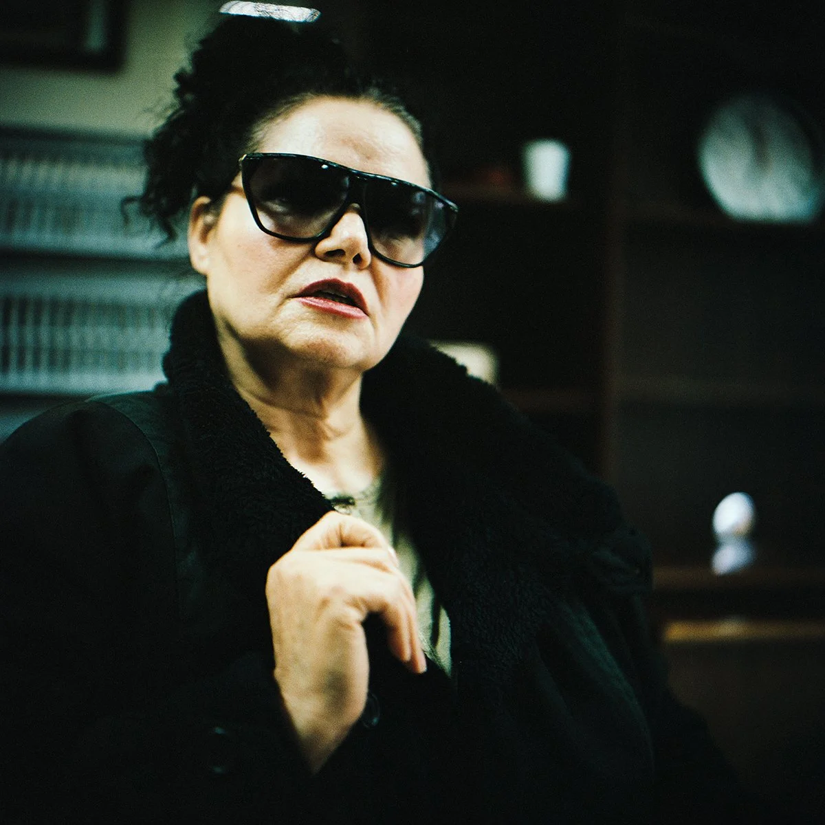 Une femme âgée portant de grandes lunettes de soleil noires et une veste noire, avec des cheveux bouclés noir foncé et un serre-tête blanc, dans un intérieur sombre.