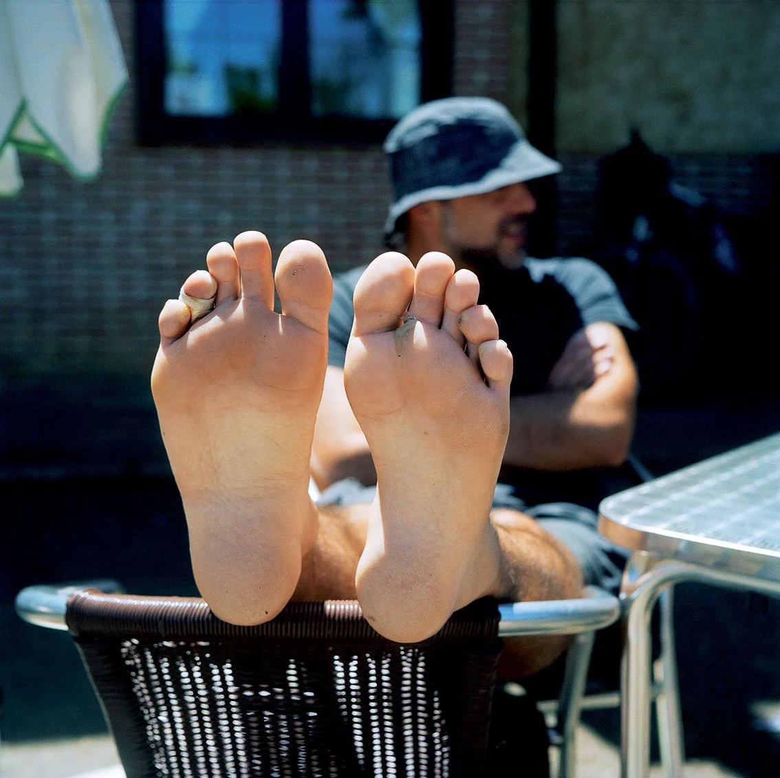 Person seated in background wearing a hat, head slightly bowed, arms crossed, sandals or bare feet extended towards foreground, on a chair, outdoors.