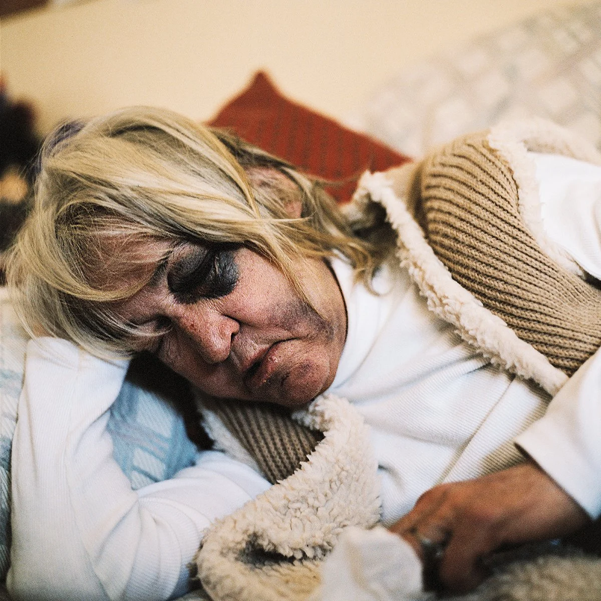 Une personne âgée avec des cheveux blonds dormir paisiblement, reposant sa tête sur un bras, avec un tissu beige à la main dans un cadre intérieur