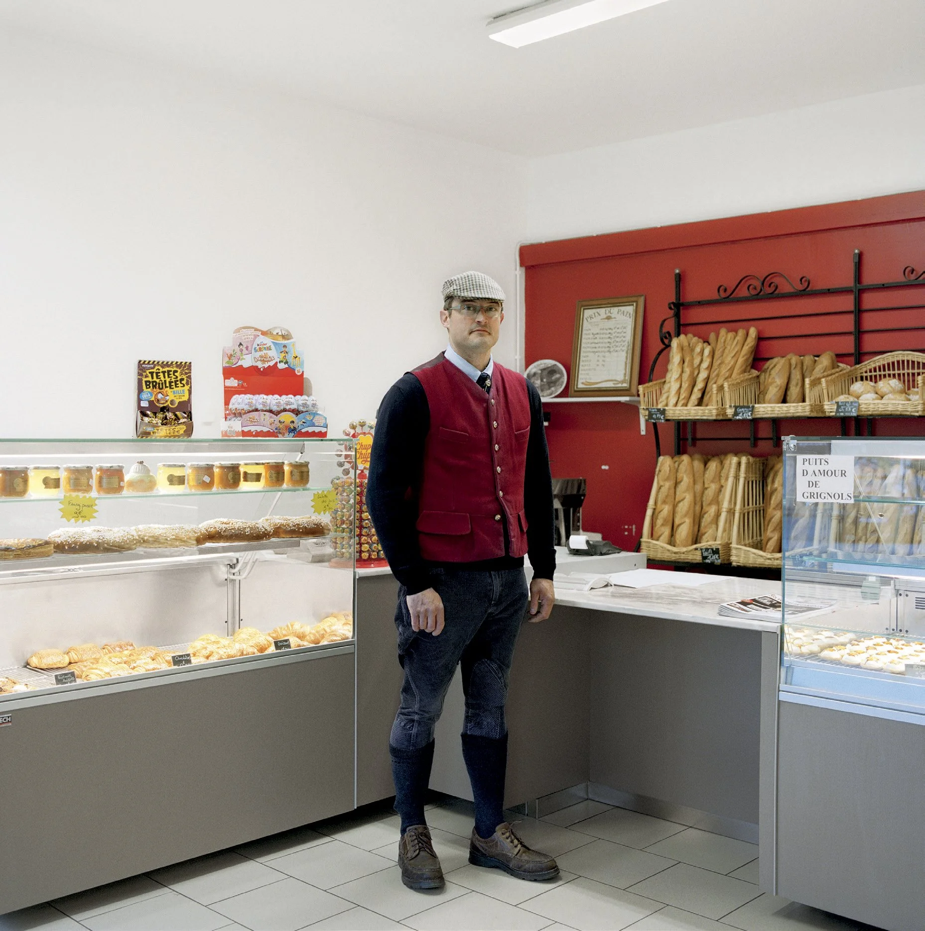 Man dressed in vintage style in a bakery, surrounded by pastries and breads, with wicker shelves and red decor in the background.