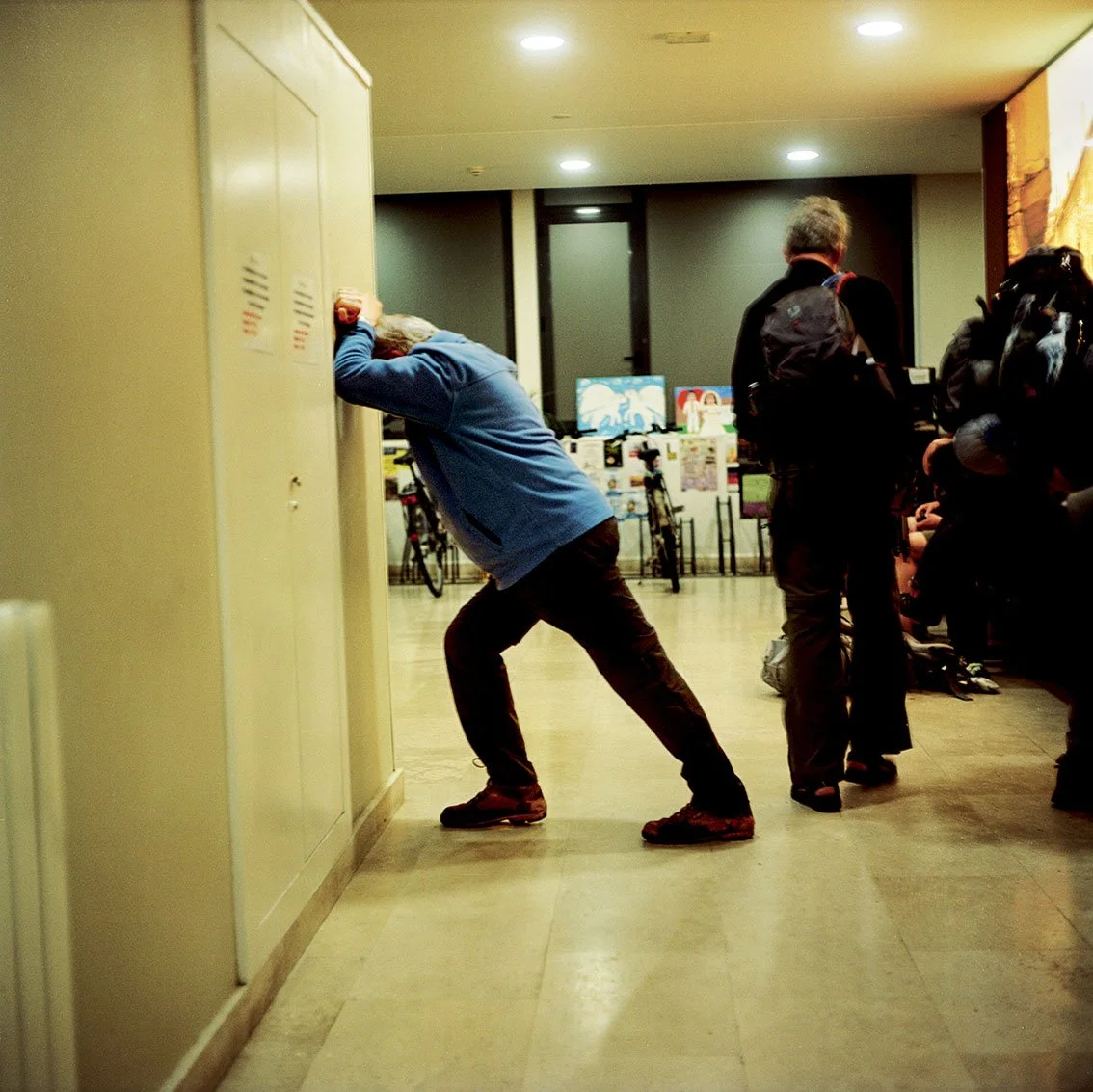 A person in a despairing position with his head against a locker, in an indoor environment with other people and screens in the background.