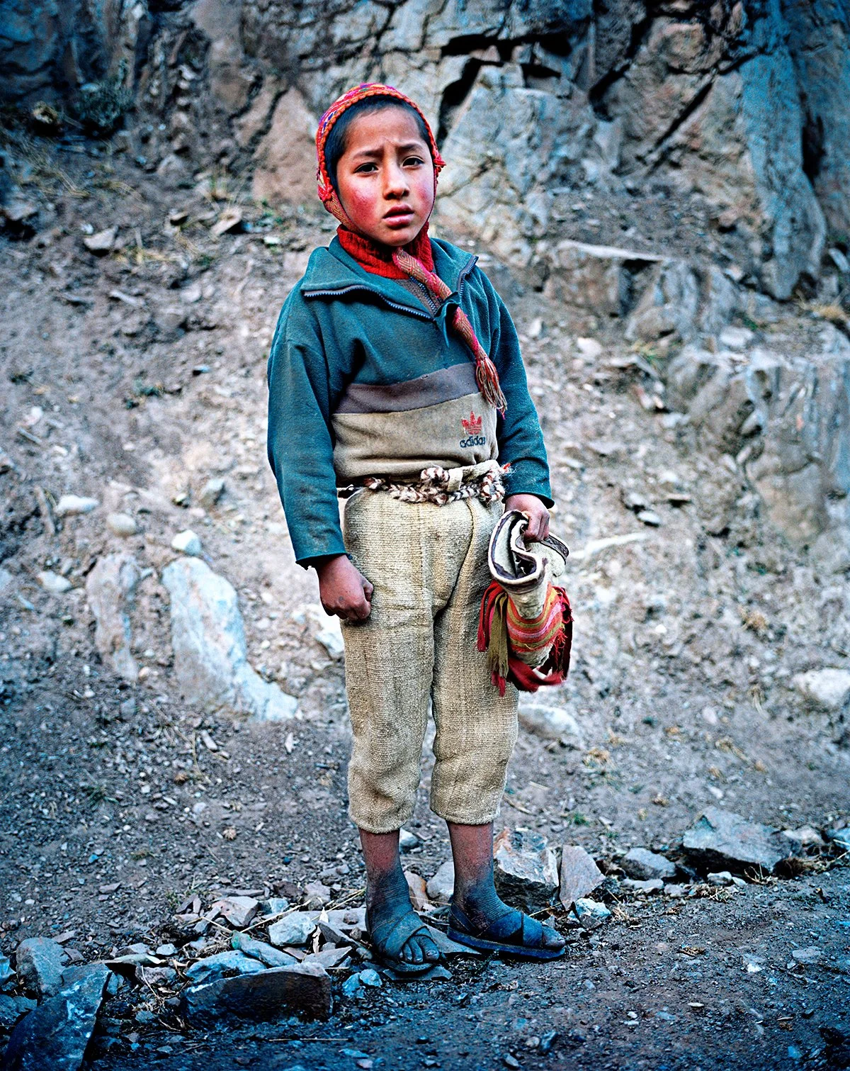 A young boy standing in a rocky environment, wearing casual clothes with a colorful cap, talking or looking at the camera.