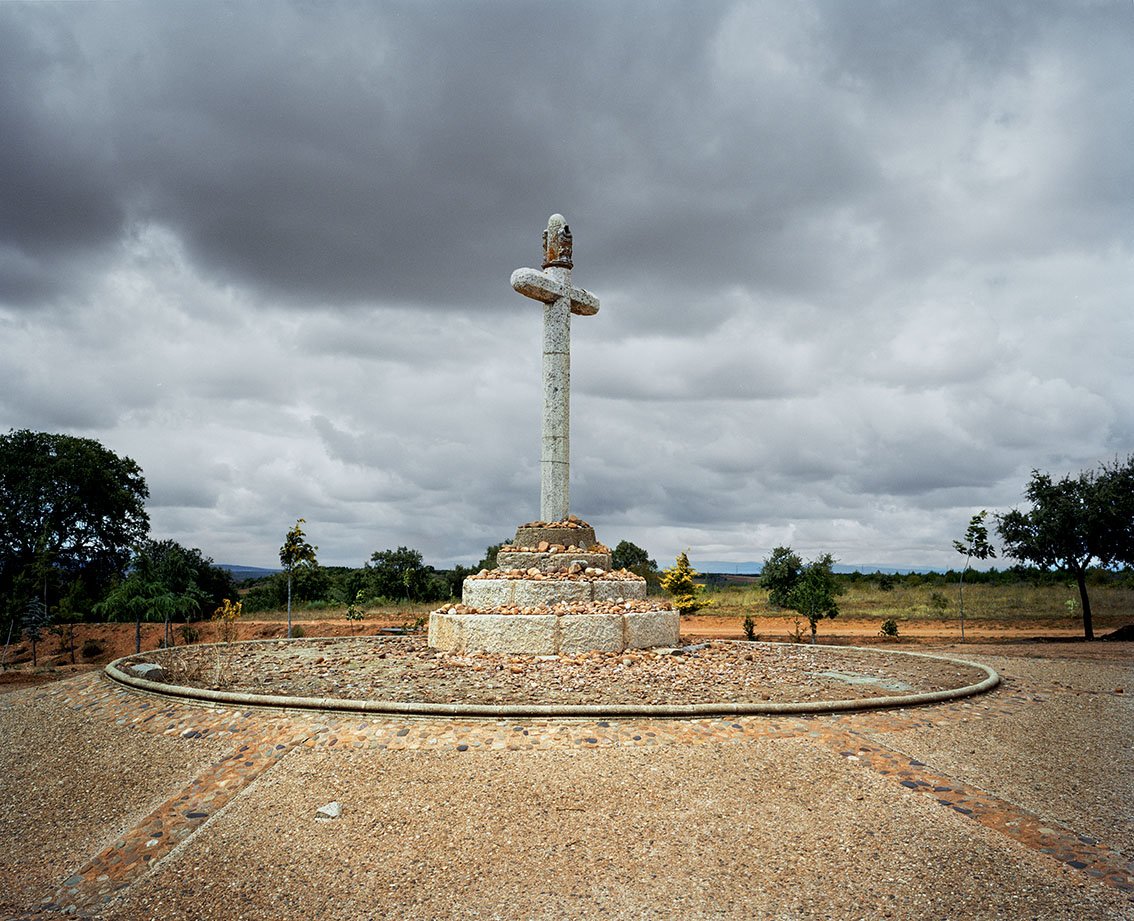 An ancient stone cross in a field under a stormy sky, surrounded by a gravel driveway.