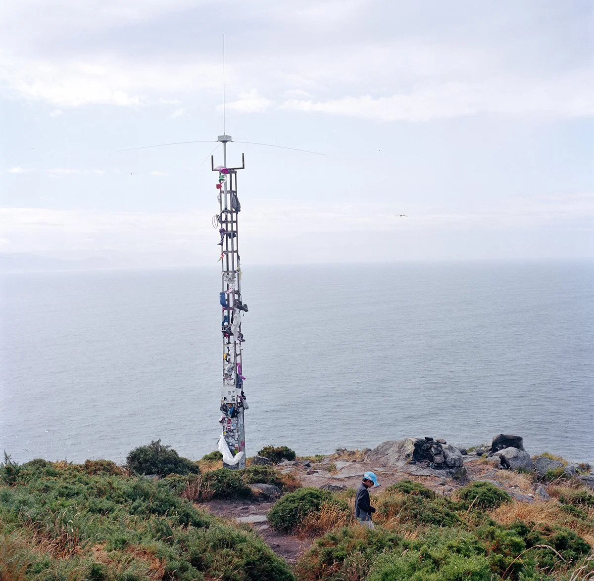 A person wearing a blue helmet walks along a path at the top of a hill with a communication tower adorned with several items of clothing and objects attached. The scene overlooks the ocean, with a cloudy sky.