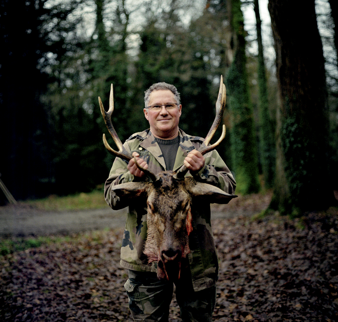 Un homme en tenue de chasse dans une forêt, tenant fièrement un cerf de large taille avec des bois impressionnants.