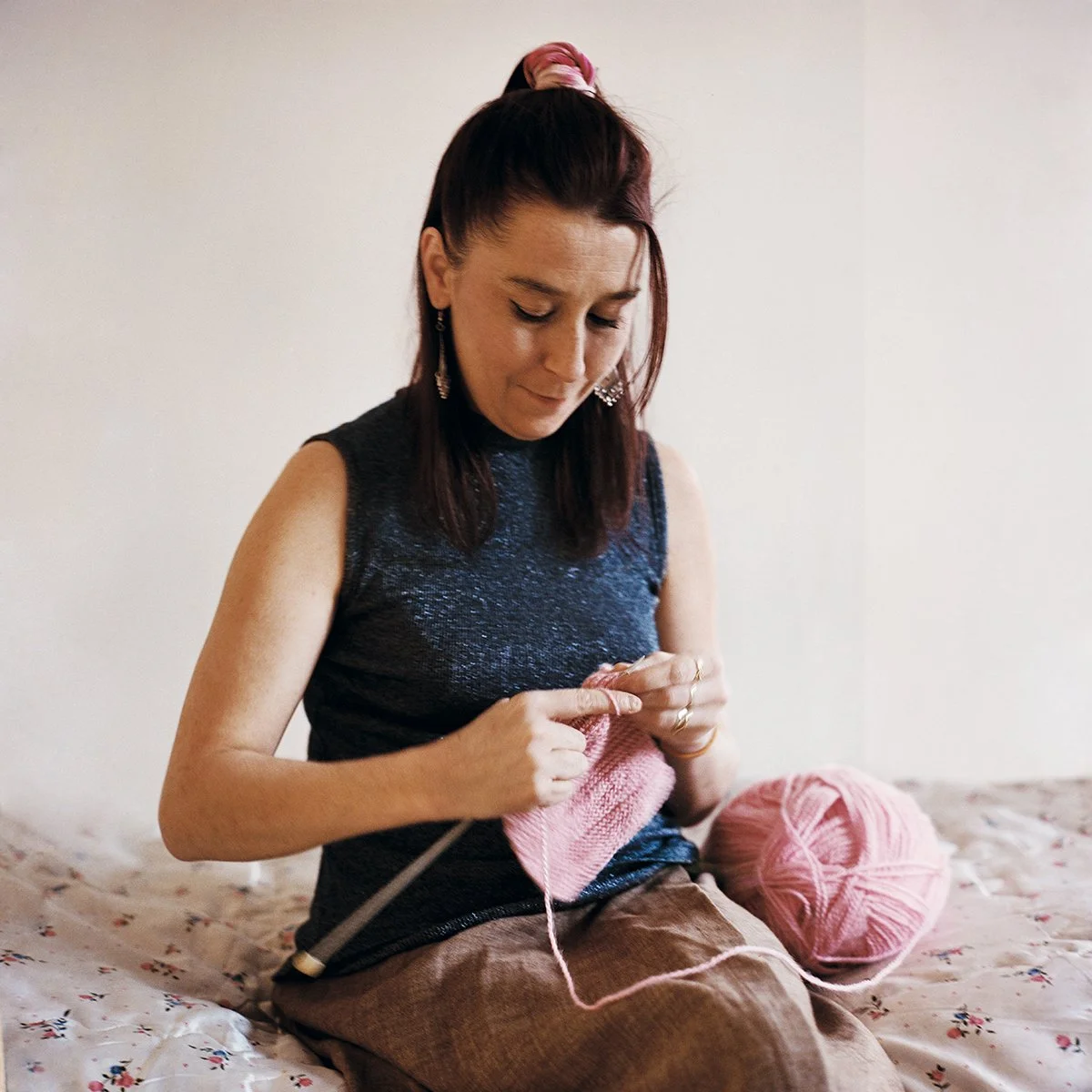 Une femme assise sur un lit en train de tricoter avec de la laine rose et des aiguilles à tricoter.