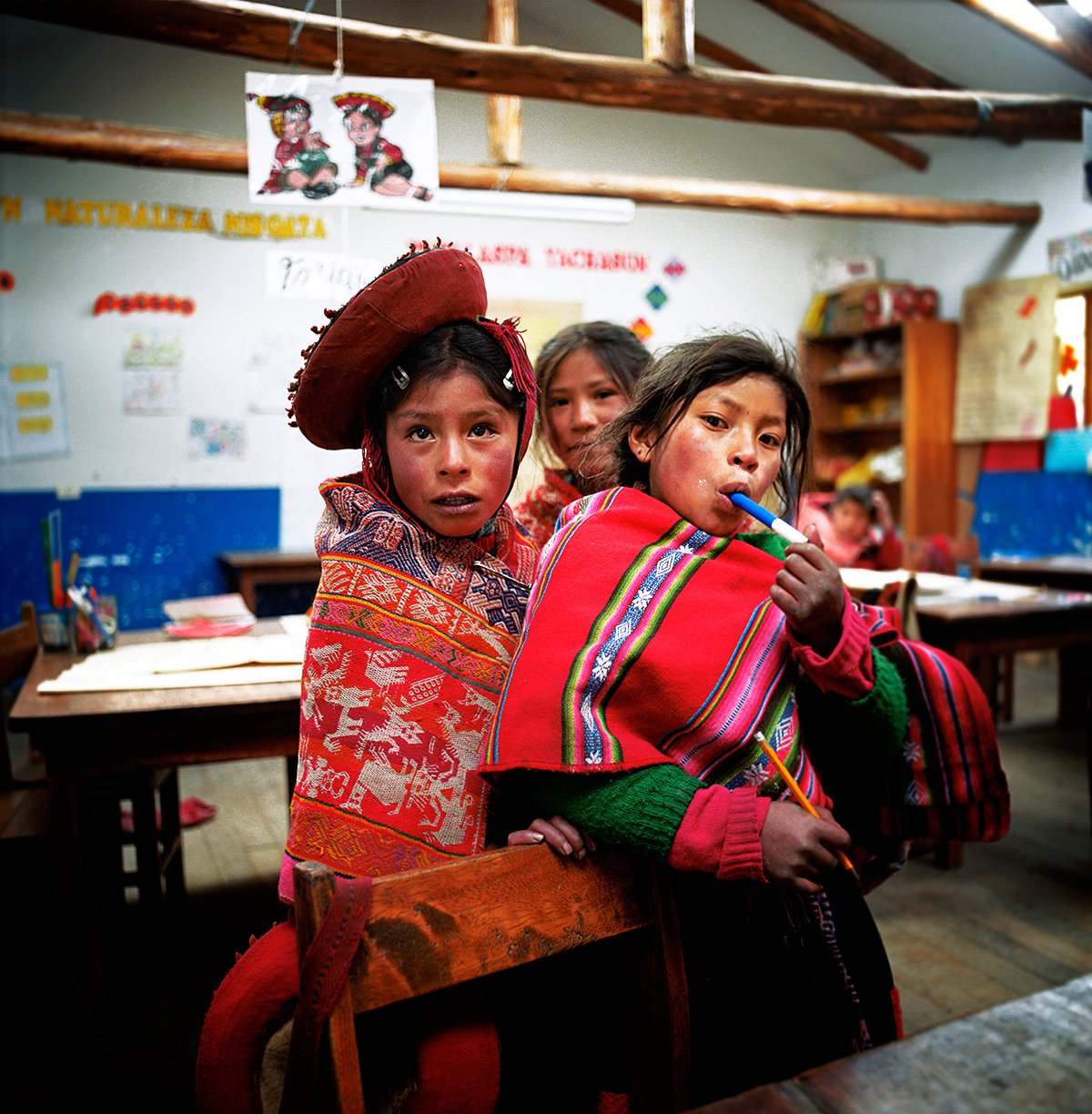 Three children in traditional dress in a school classroom, one girl holds a pen and another bites a pencil, with a chalkboard and decorations in the background.