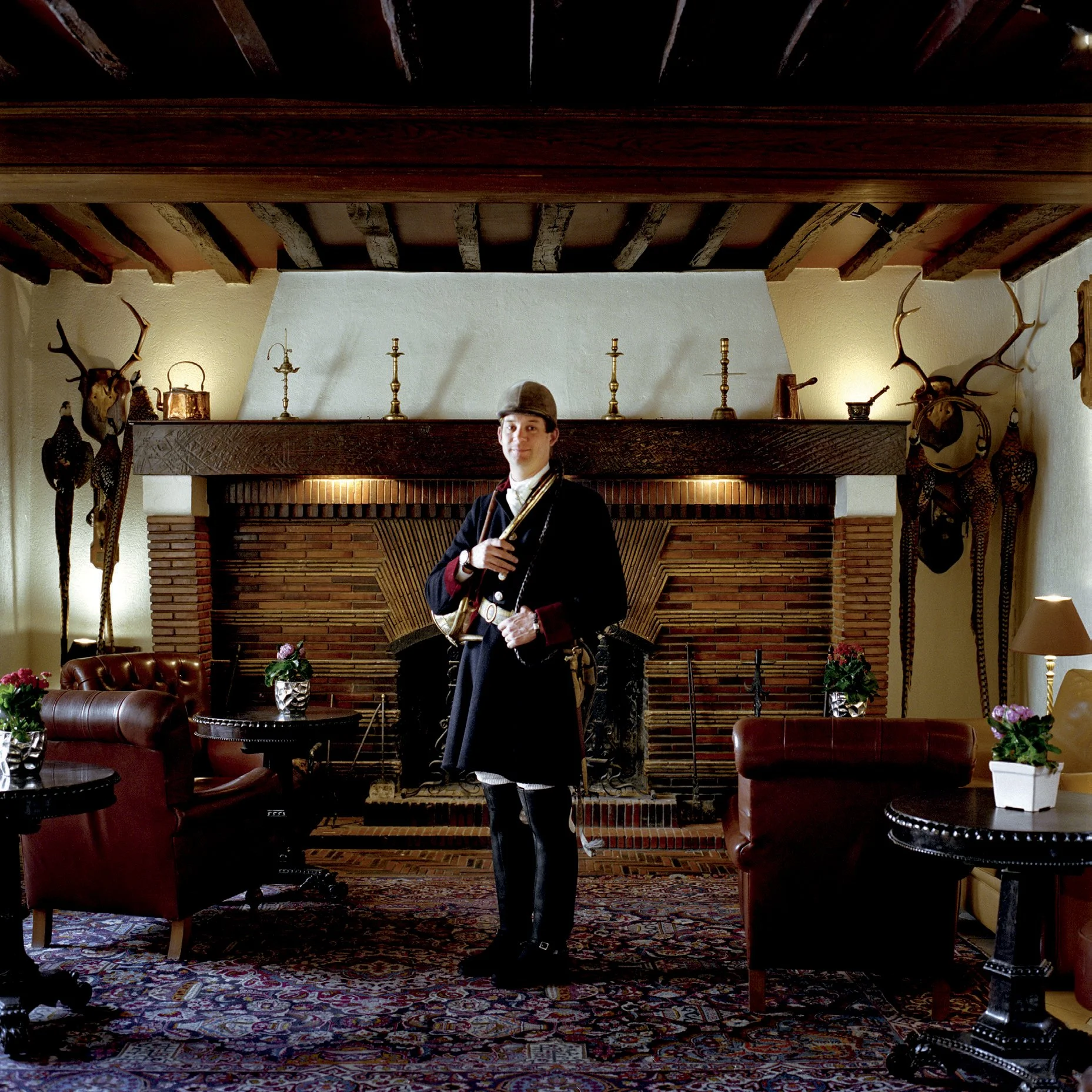A man in traditional costume in a warm room with a fireplace, decorated with deer heads and antique candlesticks.