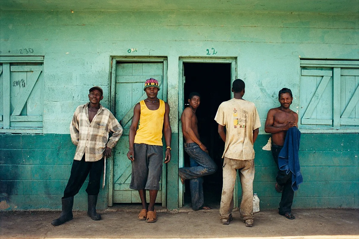 Five men standing in front of a wooden building painted turquoise green, some unclothed or in casual clothes, in an open-air environment.