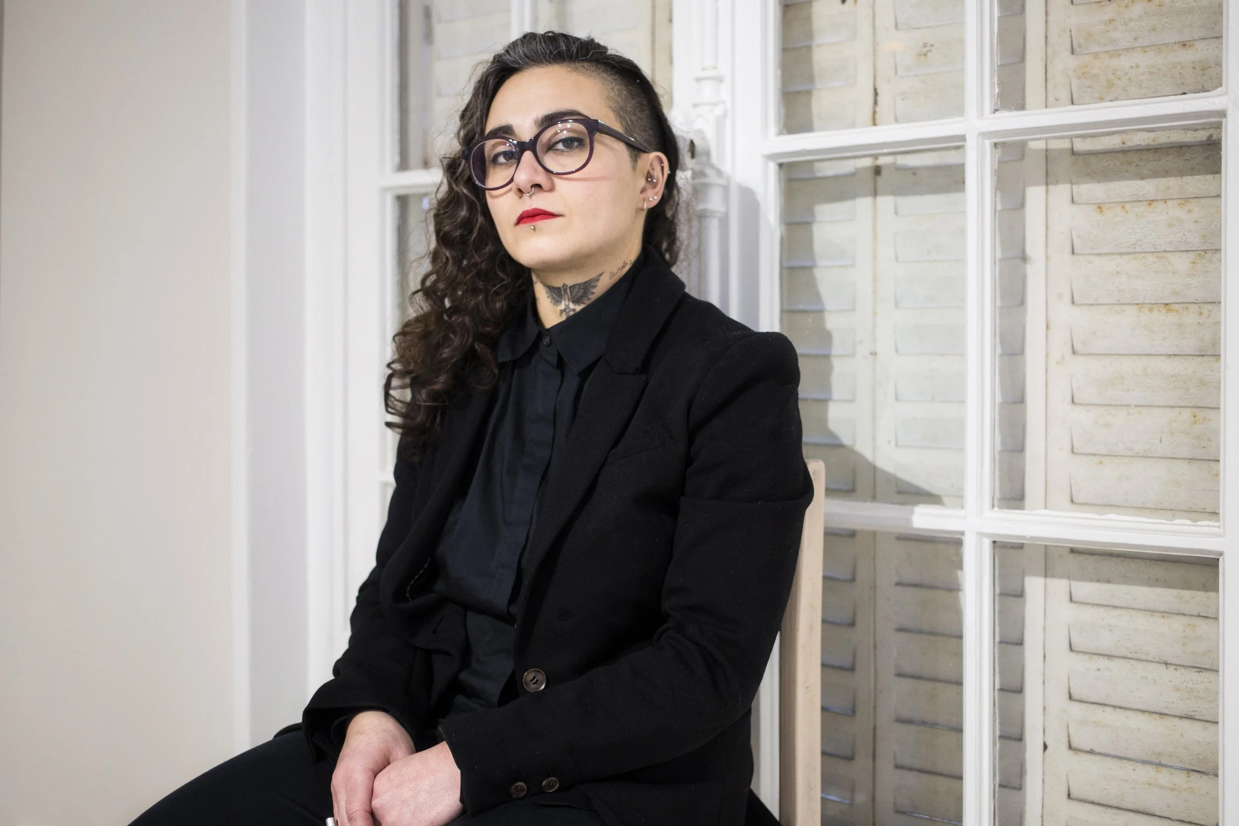 A woman with long hair and a fair complexion, wearing glasses, a black bow tie and a black jacket, seated in front of a blown-glass window.