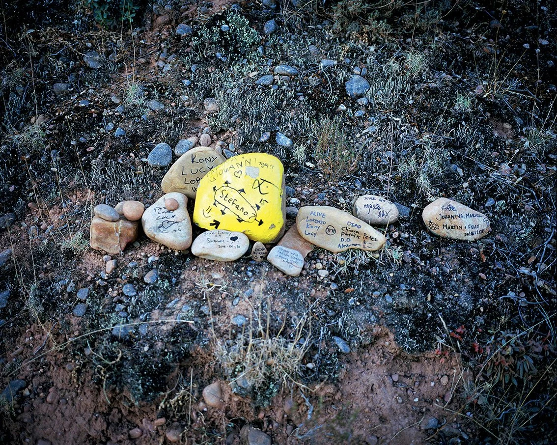 Stones engraved with messages and drawings, arranged in a circle on the dry earth floor, in a natural environment with small plants and pebbles.