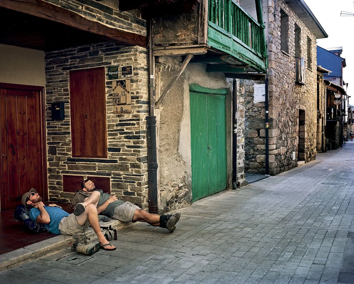 Two men lying on the sidewalk in front of a stone house with a wooden door and wall, on a cobblestone street, under a wooden balcony painted green.