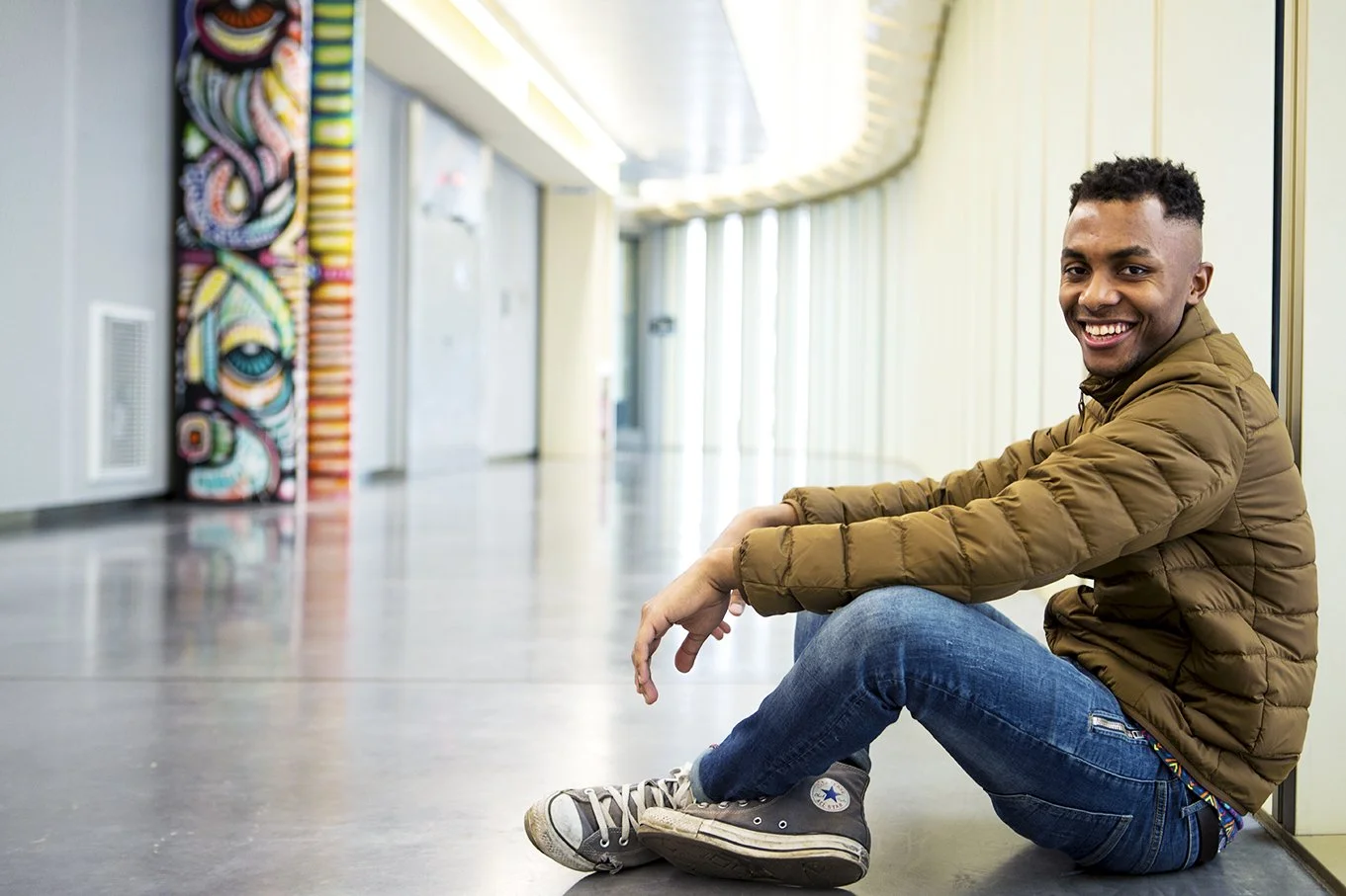 A smiling young man sits on the floor of a bright corridor, wearing a brown jacket, jeans and grey sneakers. To his left, a wall decorated with a colorful work of art.