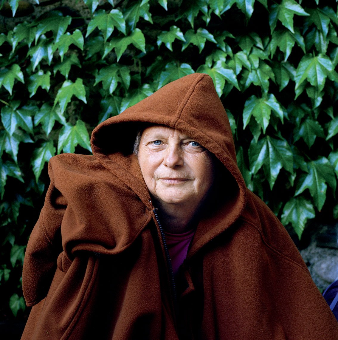 An elderly woman wearing a brown hooded coat, seated in front of a wall of green ivy leaves.