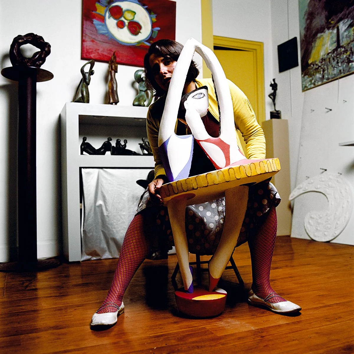 Woman with an artistic sculpture in the center, in an interior decorated with works of art and statues. The walls feature colorful paintings.