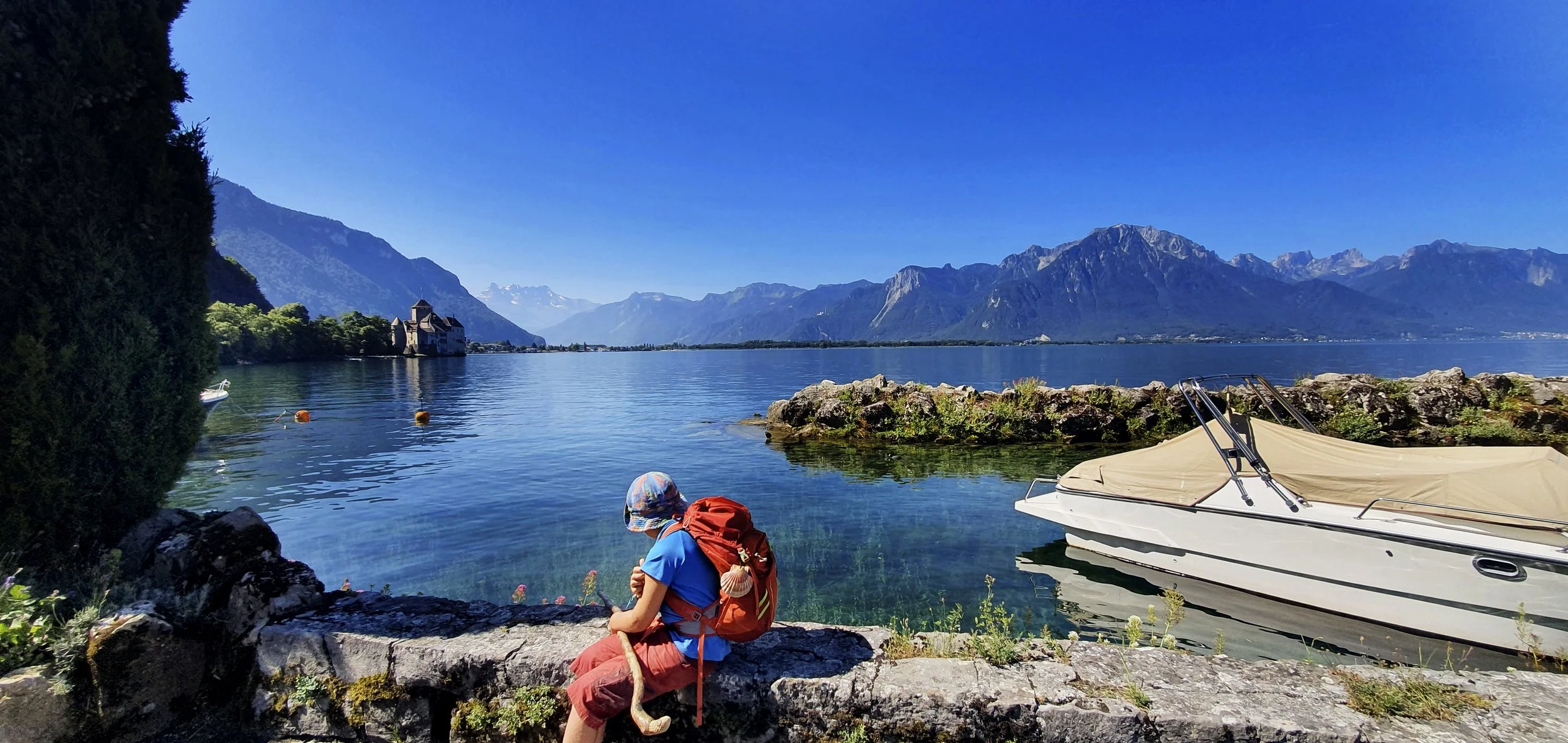 Young child with red backpack, hat, displaying heat stick, sitting on a stone wall, near a lake surrounded by towering mountains under a clear sky.