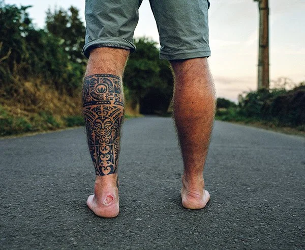 Personne marchant pieds nus sur une route, avec une jambe tatouée et l'autre nue, dans un environnement extérieur au crépuscule.