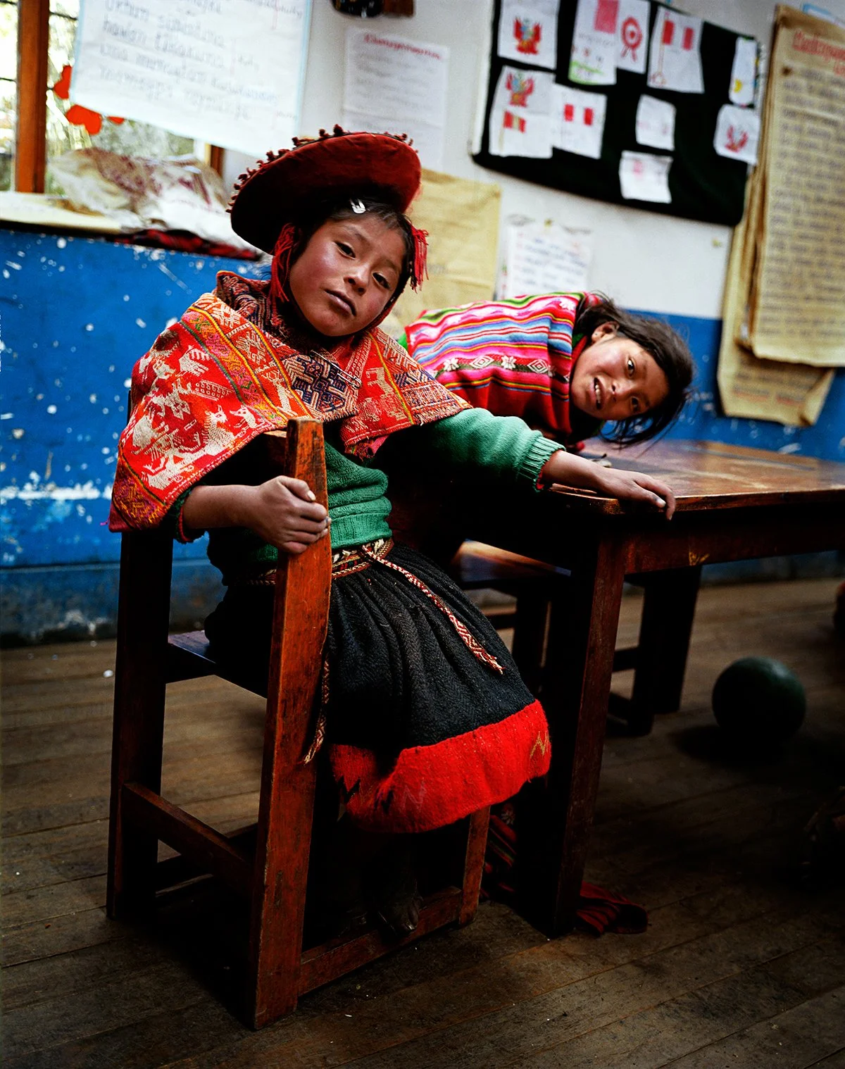 Two children in traditional Andean costumes sit in a colorful room, one seated on a wooden chair with a red hat, the other leaning over a table, decorated with posters and papers on the wall.