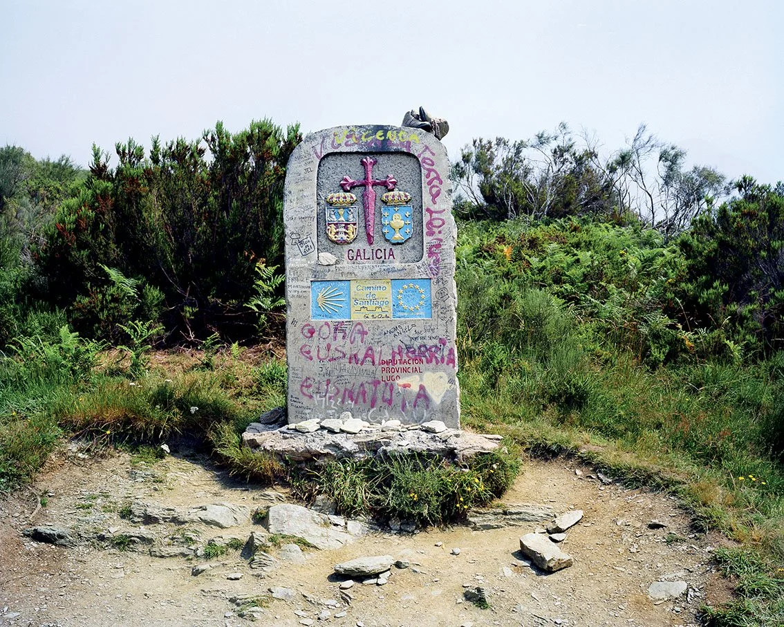 Stele marked with decorations, graffiti and the words 'Galicia', located in a natural area with bushes.