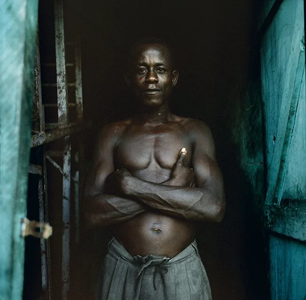 Homme debout dans un espace sombre, poitrine nue, bras croisés, regardant vers l'avant.