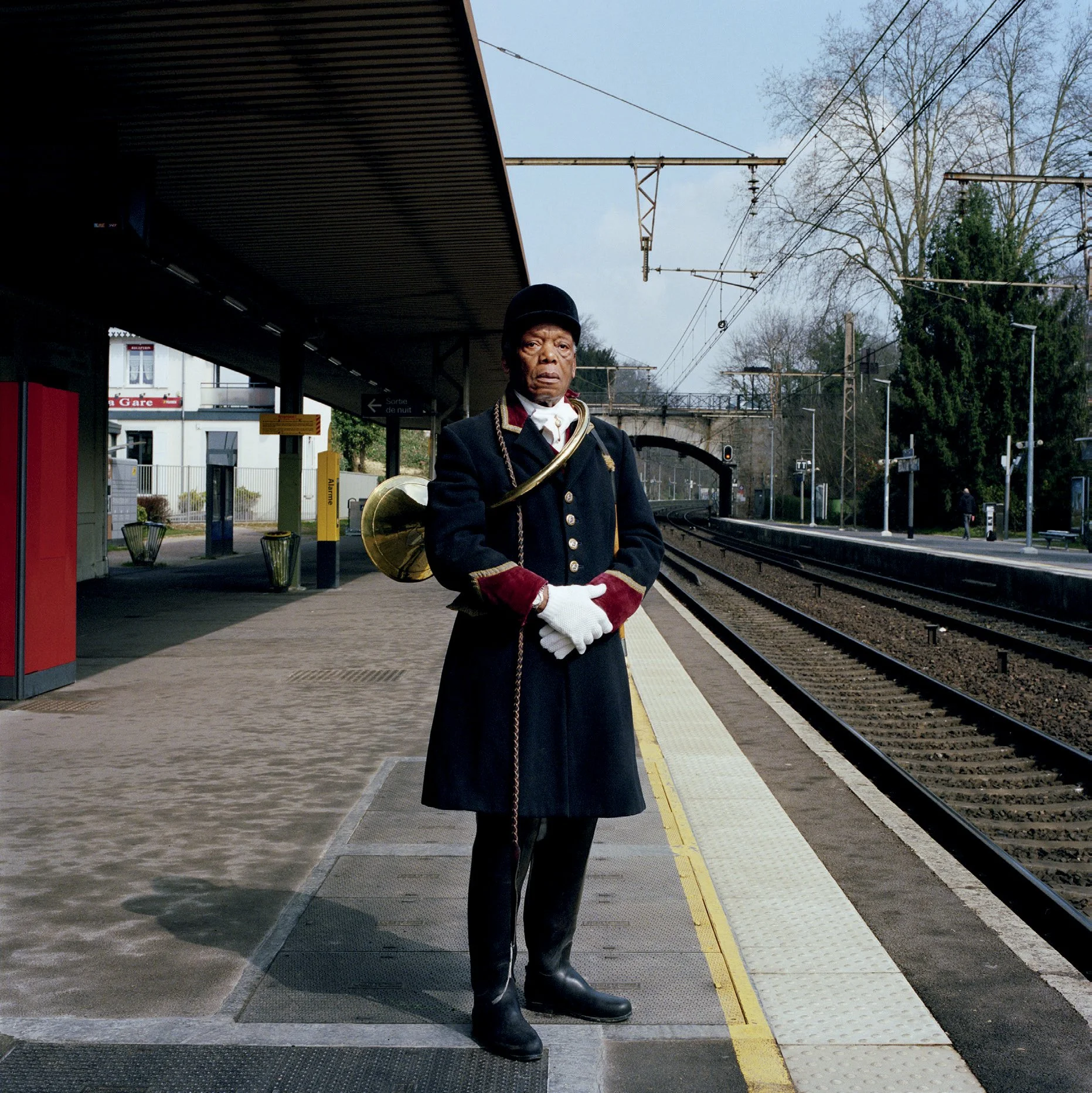 A man in a border guard uniform with helmet and black uniform with red details, holding a trumpet, at a train station. The background shows rails, trees and a tunnel. It's an outdoor scene on a clear day.