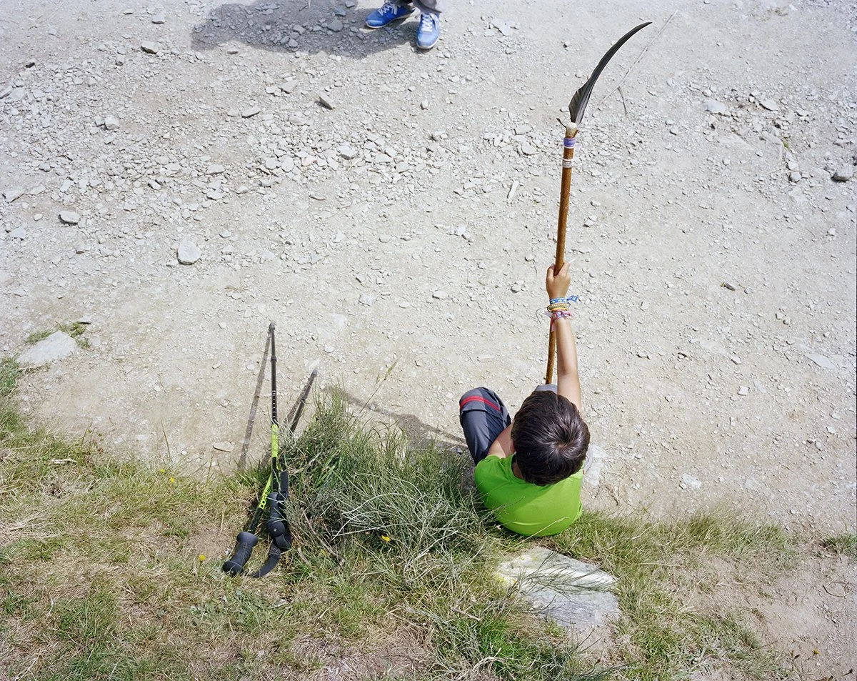 A child sitting on a rock ledge, looking down, holding a spear or wooden weapon with a stone at its end. Sticks or hiking poles lie in the grass beside him.