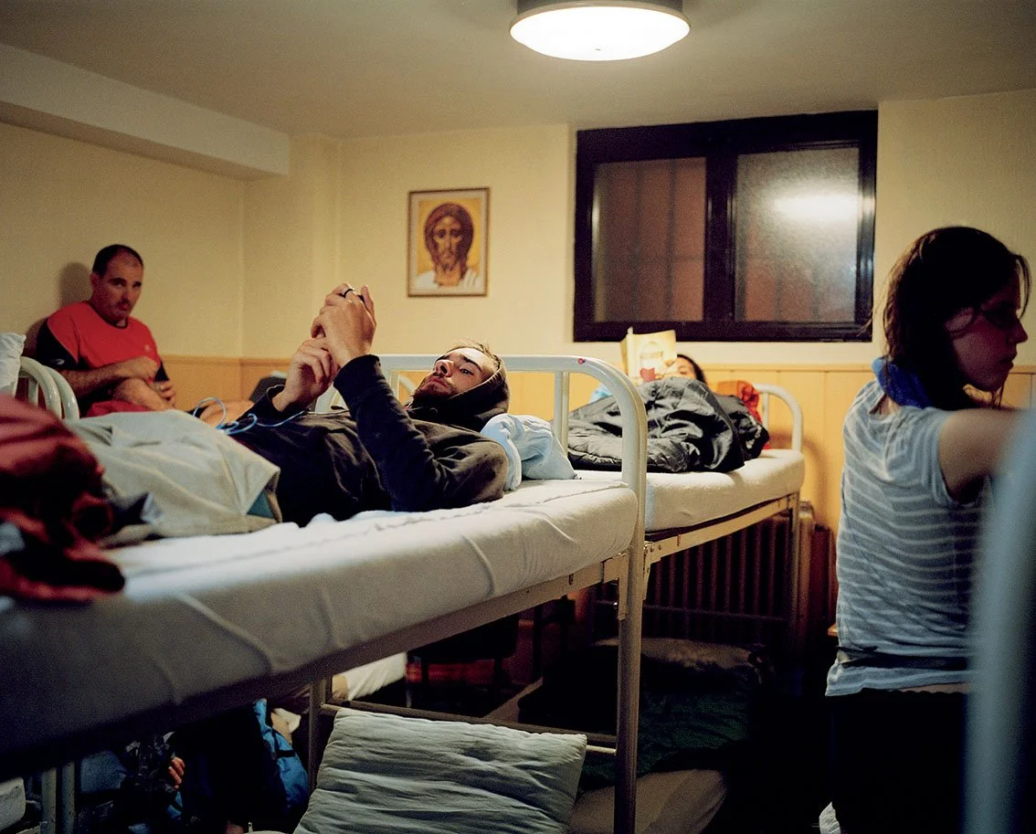 Group of young adults in a dormitory room, some lying in bunk beds and others sitting, looking at their phones, with a religious icon painted on the wall.