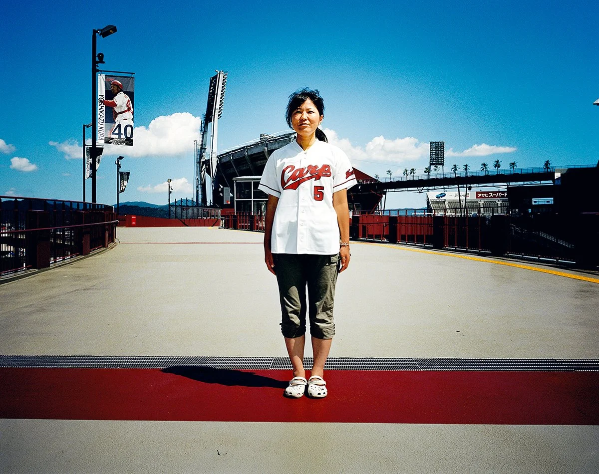 Une femme portant un maillot de baseball blanc avec le mot « Care » en rouge, numéro 5, se tient sur un pont à l'extérieur d'un stade de baseball. Le ciel est ensoleillé avec quelques nuages, et il y a des panneaux publicitaires et des lampadaires en
