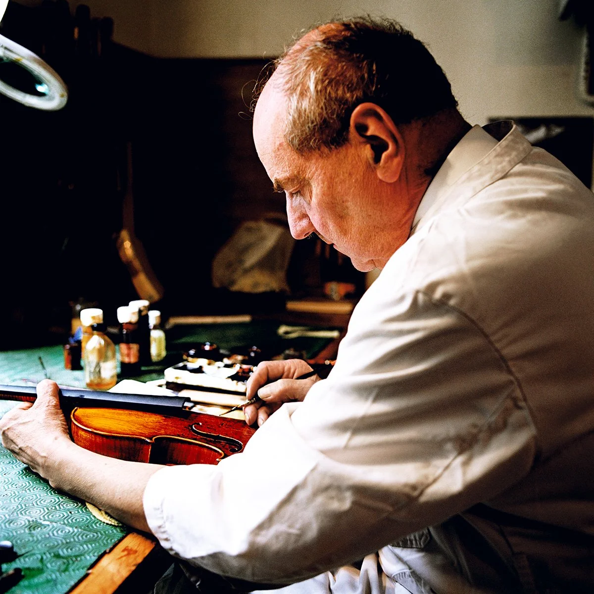 A man in a white coat repairing a violin in a workshop, with small bottles and tools on the table.