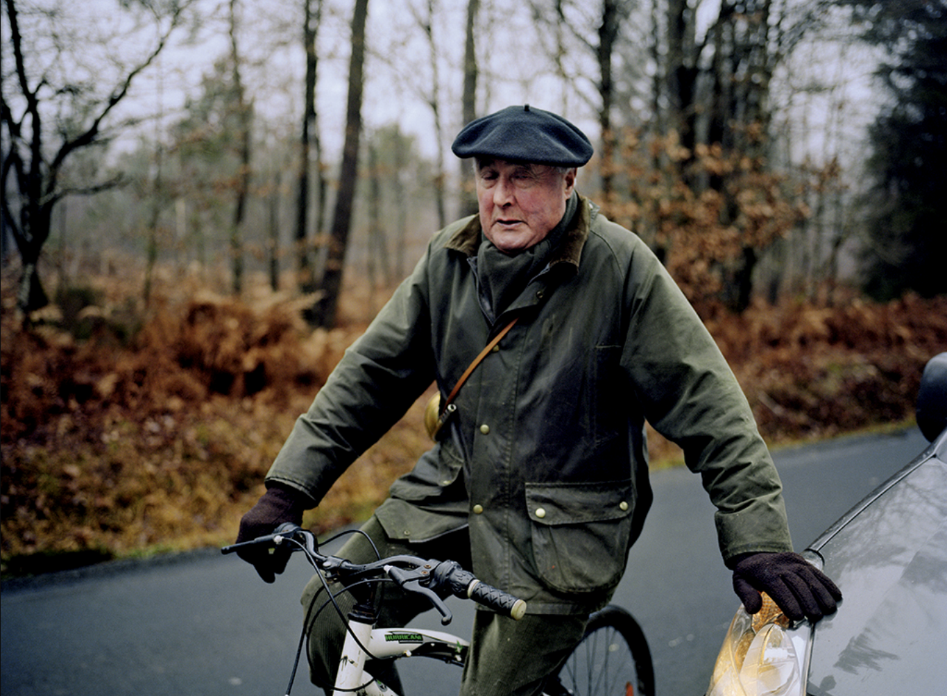 Un homme âgé portant un manteau vert, un béret noir et des gants marron, fait du vélo dans une forêt en automne.