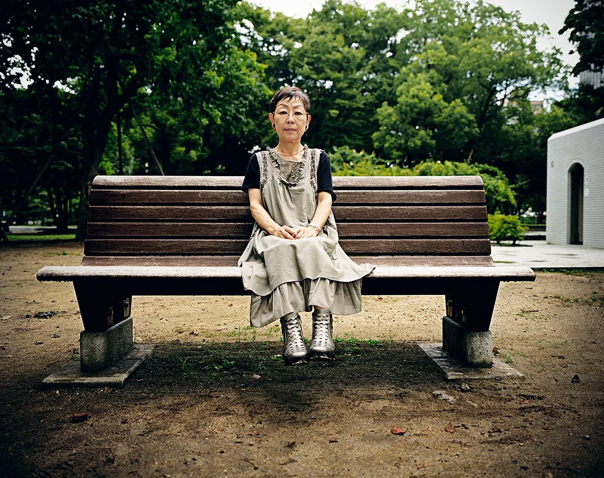 Une femme âgée assise seule sur un banc en bois dans un parc vert, portant une robe beige et des bottes en métal.